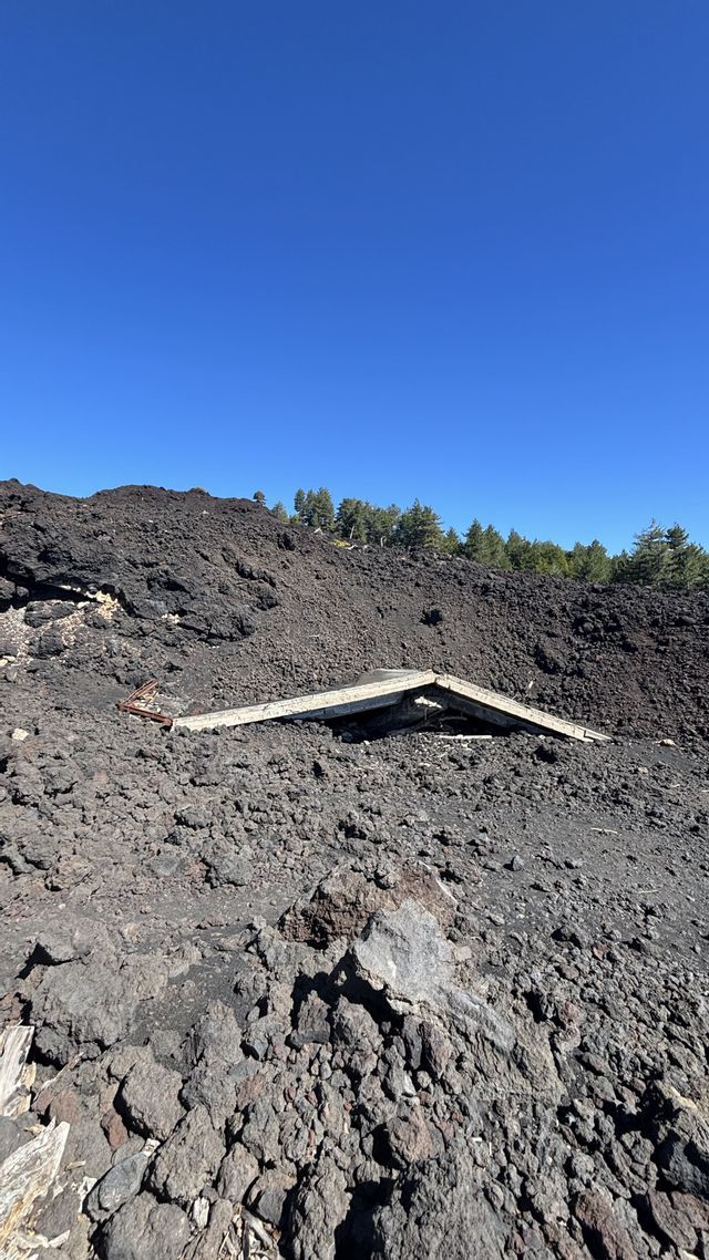 Il tetto di un edificio è quasi completamente sepolto da un campo di roccia vulcanica nera, con pini sullo sfondo sotto un cielo azzurro e limpido.