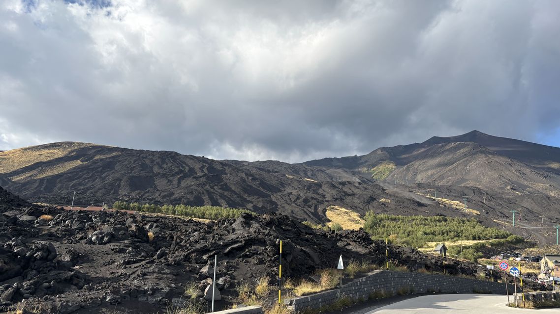 Un vasto paesaggio vulcanico con scuri pendii di roccia lavica, chiazze di erba secca e una strada in primo piano sotto un cielo nuvoloso.