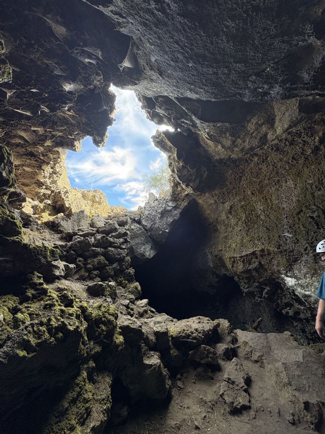 Vista dall'interno di una buia grotta rocciosa e muschiosa, che si apre su un cielo azzurro con nuvole bianche.