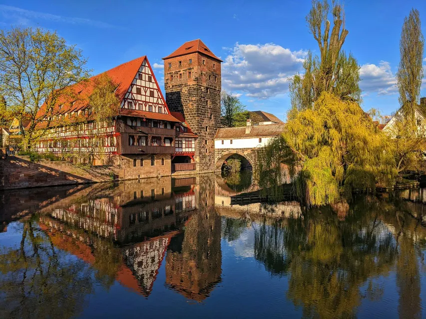 Edifici medievali a graticcio e una torre di pietra perfettamente riflessi in un fiume calmo sotto un cielo azzurro.