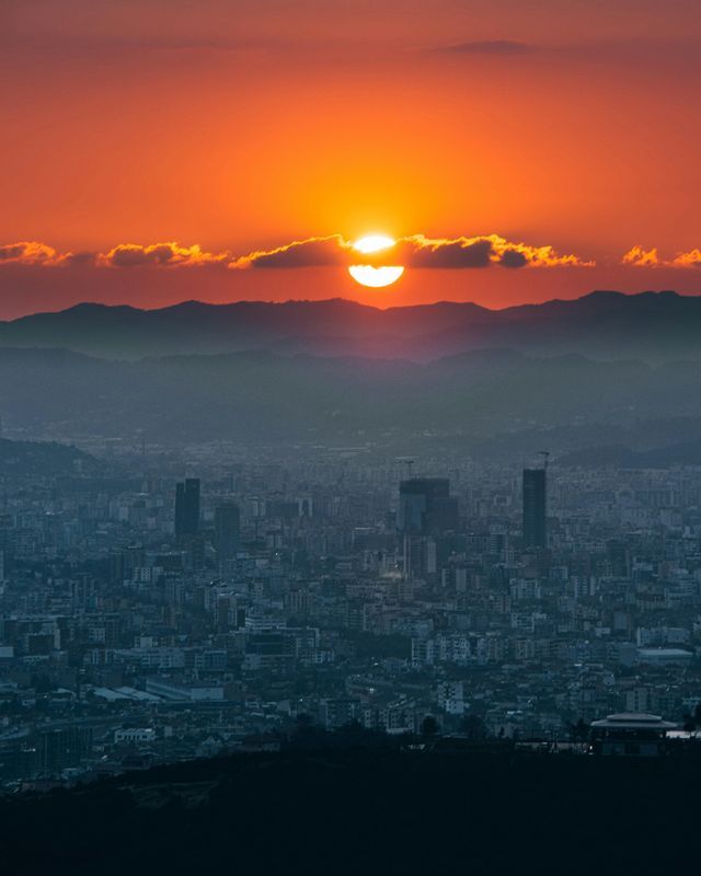 Un sole arancione intenso tramonta dietro una lontana catena montuosa, che si affaccia su una vasta città avvolta nella foschia.