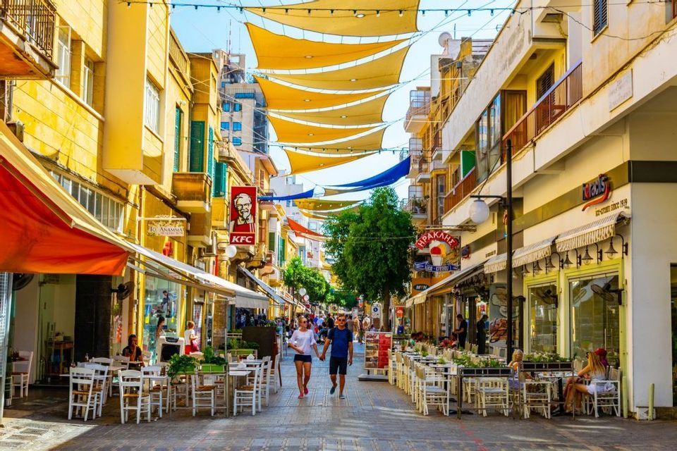 Un couple se promène dans une rue piétonne animée bordée de cafés et de boutiques, sous de grands stores de soleil jaunes suspendus entre les bâtiments.