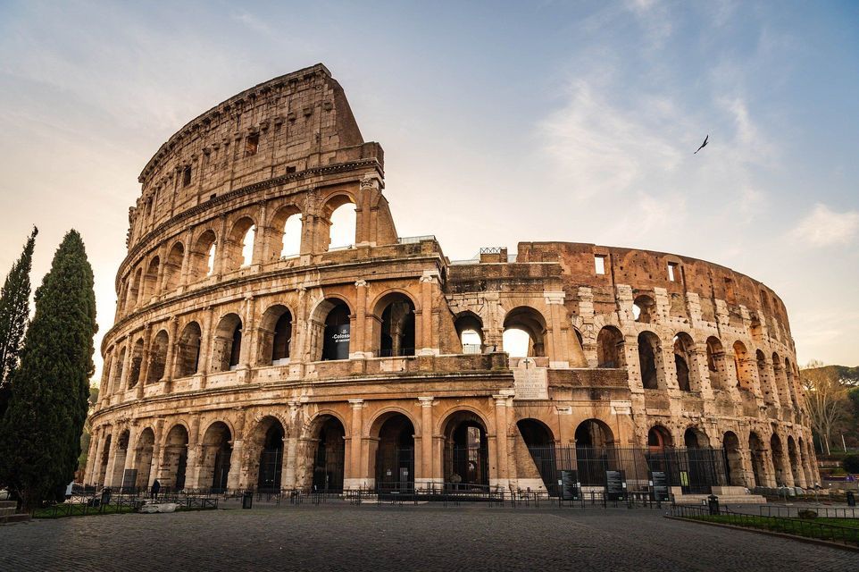 El antiguo anfiteatro Coliseo en Roma, visto desde una plaza de adoquines vacía al amanecer bajo un cielo parcialmente nublado.