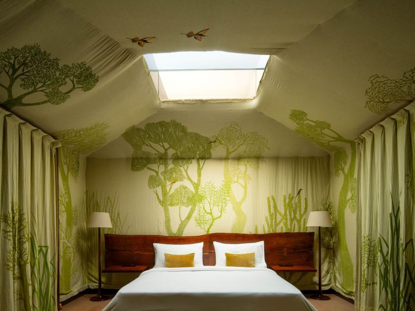Ein Doppelbett mit einem Holzkopfteil in einem zeltähnlichen Zimmer mit Stoffwänden, grünen Baumillustrationen und einem Oberlicht.