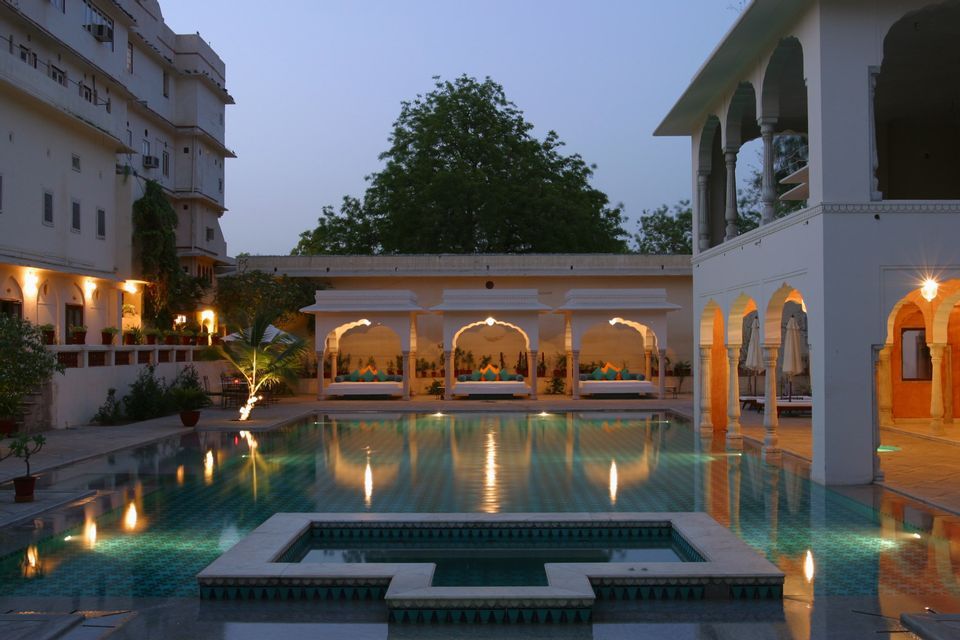 Una piscina illuminata nel cortile di un hotel al tramonto, con luci che si riflettono sull'acqua e cabana ad arco sullo sfondo.