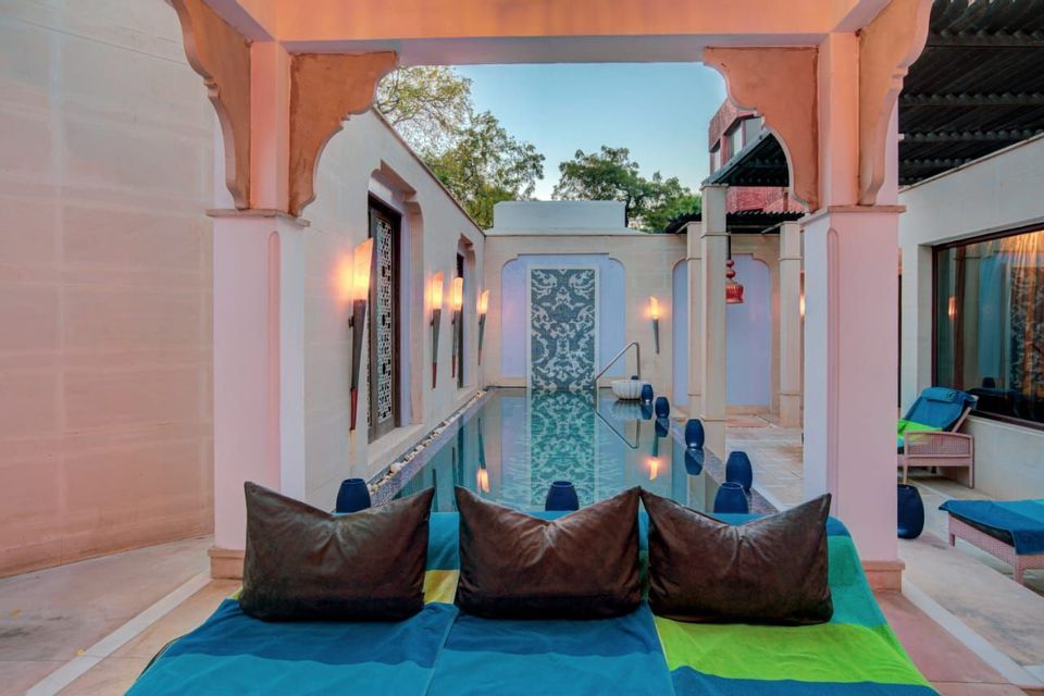 Bunte Liegestühle mit dunklen Kissen stehen an einem schmalen Pool in einem eleganten Innenhof mit beleuchteten Wandlampen.