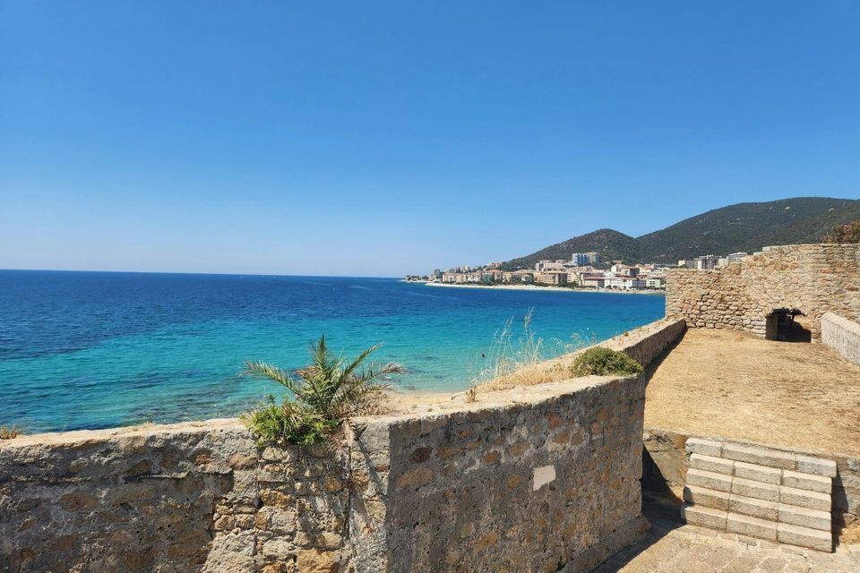Une vue depuis un mur de pierre surplombant une ville côtière sur une baie aux eaux turquoise et aux collines verdoyantes sous un ciel clair.