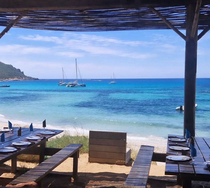 Deux tables de pique-nique en bois dressées pour un repas sur une plage de sable, surplombant une mer turquoise avec plusieurs voiliers.