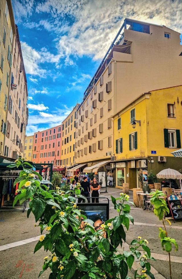Une rue étroite et sinueuse bordée de bâtiments colorés sous un ciel bleu partiellement nuageux, avec des gens marchant sur le trottoir.