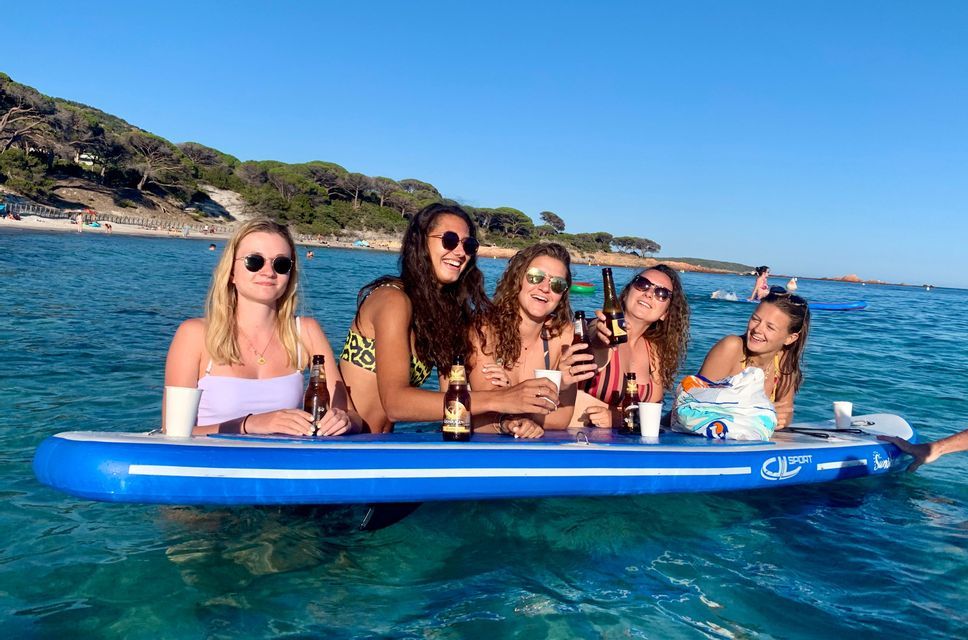 Un groupe WeRoad de cinq femmes en bikini, souriantes et buvant des bières autour d'un paddleboard en mer.
