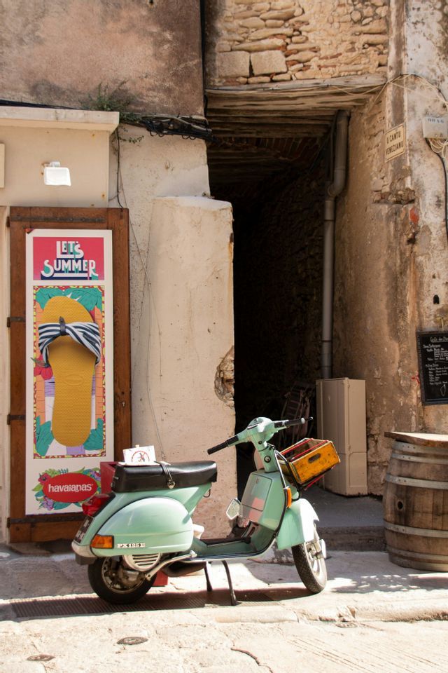 Un scooter vintage vert clair avec une caisse jaune est garé dans une étroite ruelle en pierre à côté d'une publicité estivale.