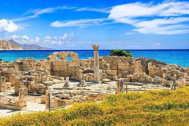 Anciennes ruines de pierre avec une colonne dressée, surplombant une mer d'un bleu éclatant, et des fleurs jaunes au premier plan.