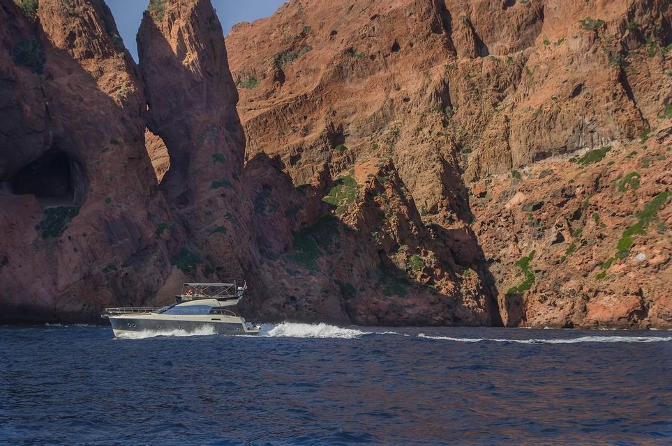 Un yacht à moteur navigue sur la mer d'un bleu profond le long d'une côte spectaculaire de falaises de roche rouge avec une arche naturelle.