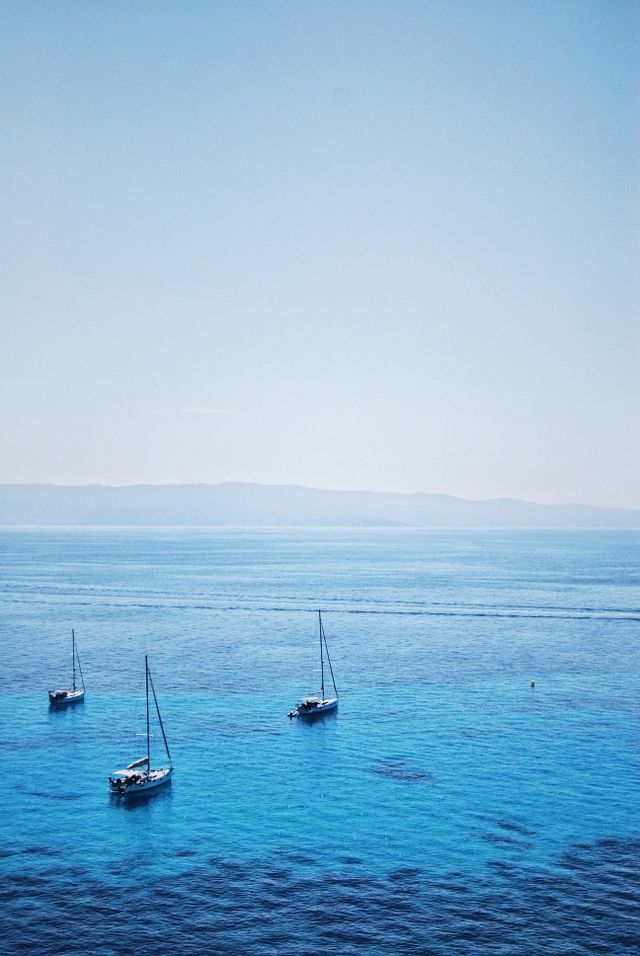Trois voiliers flottent sur une eau calme et turquoise près d'un rivage lointain sous un ciel clair.