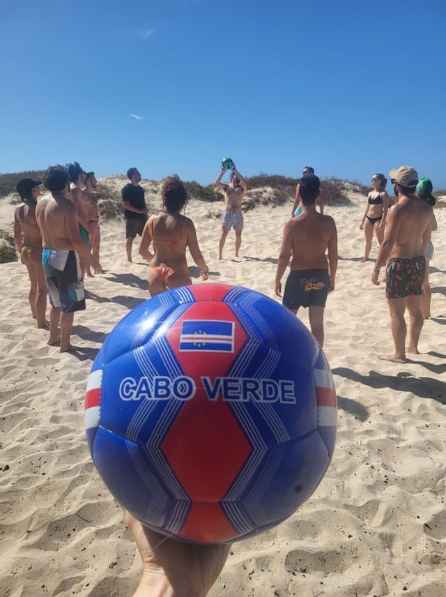 Una mano sostiene un balón de fútbol azul y rojo de 'Cabo Verde' en primer plano, mientras un grupo de WeRoad juega en una playa de arena al fondo.