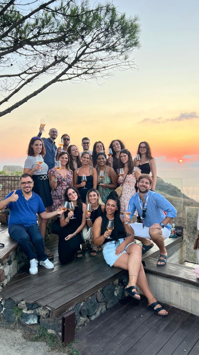 Un gruppo WeRoad in posa per una foto su una terrazza, con calici di vino, durante un tramonto dorato sul mare.