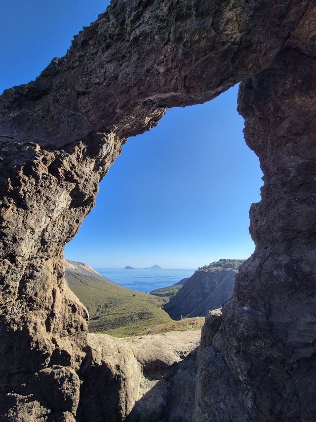Un paesaggio costiero con verdi colline e isole lontane, ammirato attraverso un arco di roccia naturale in una giornata di sole.