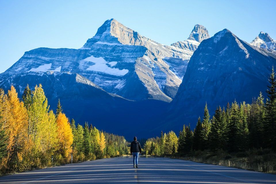 Una persona cammina al centro di una strada vuota che conduce verso grandi montagne innevate e foreste autunnali.