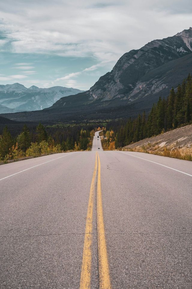Una strada asfaltata a due corsie con linee gialle si estende attraverso una foresta autunnale verso una vasta catena montuosa in lontananza.