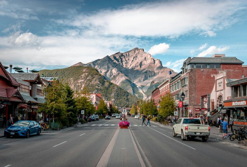 Una strada ampia attraversa una città in direzione di un'imponente vetta rocciosa, sotto un cielo parzialmente nuvoloso.
