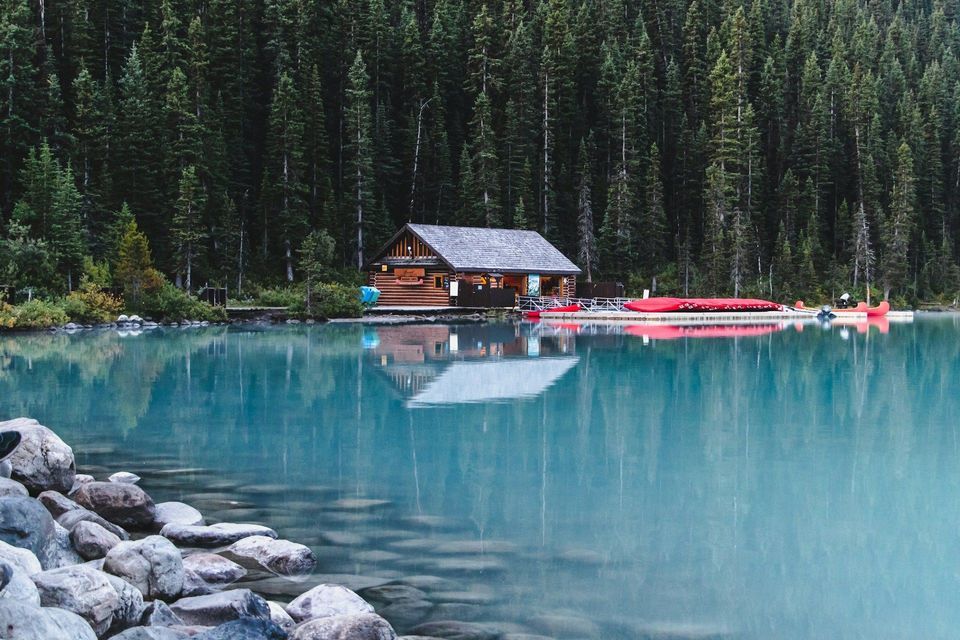 Una baita di legno con canoe rosse ormeggiate sulla riva di un lago calmo e turchese, con alle spalle una fitta pineta.