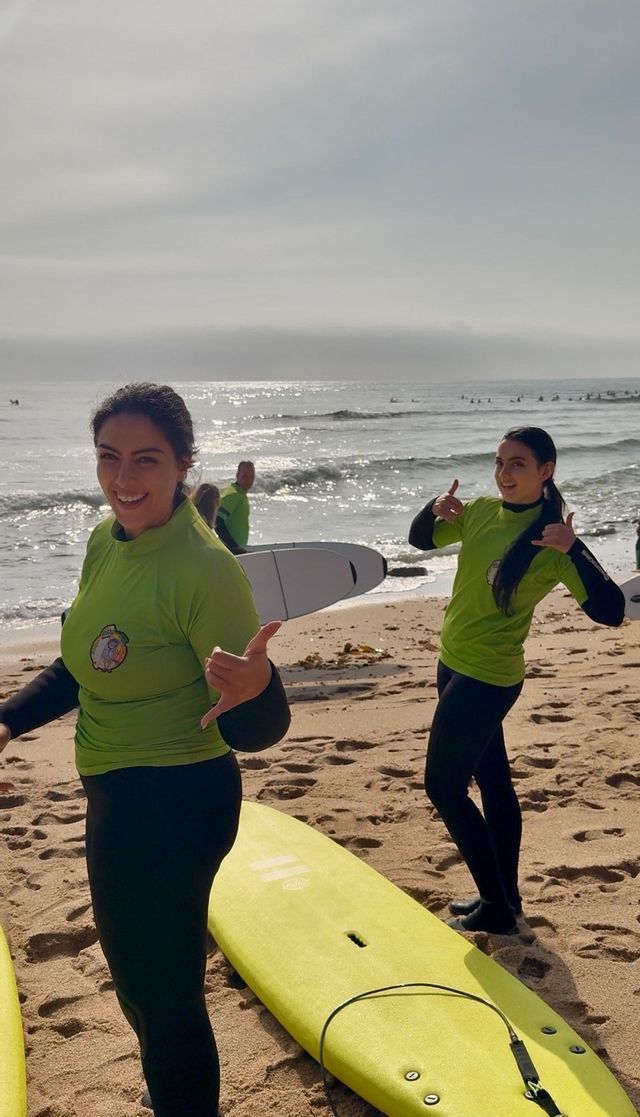 Due donne in viaggio di gruppo WeRoad, con magliette da surf verdi e mute, sorridono con le loro tavole da surf su una spiaggia sabbiosa.