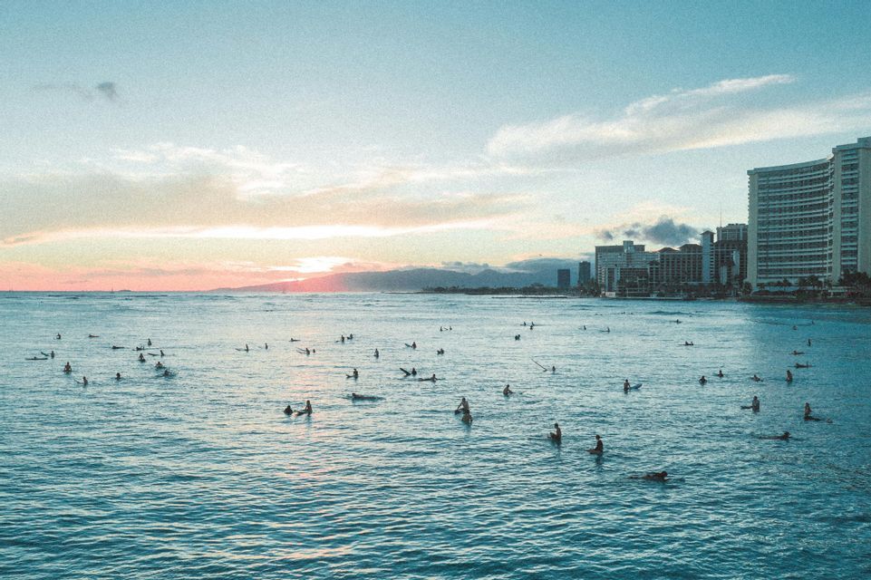 Molti surfisti sulle loro tavole sono sparsi nell'oceano vicino a uno skyline cittadino al tramonto.