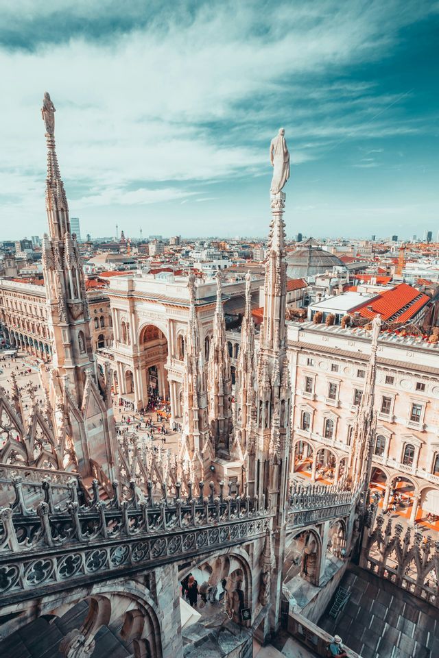 Ornate spires on a gothic cathedral rooftop overlook a bustling city square and buildings under a partly cloudy sky.