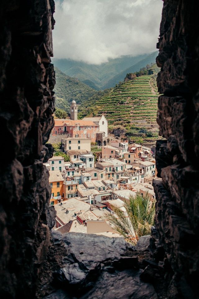 A view through a stone archway of a colorful hillside village with a church and terraced vineyards in the background.