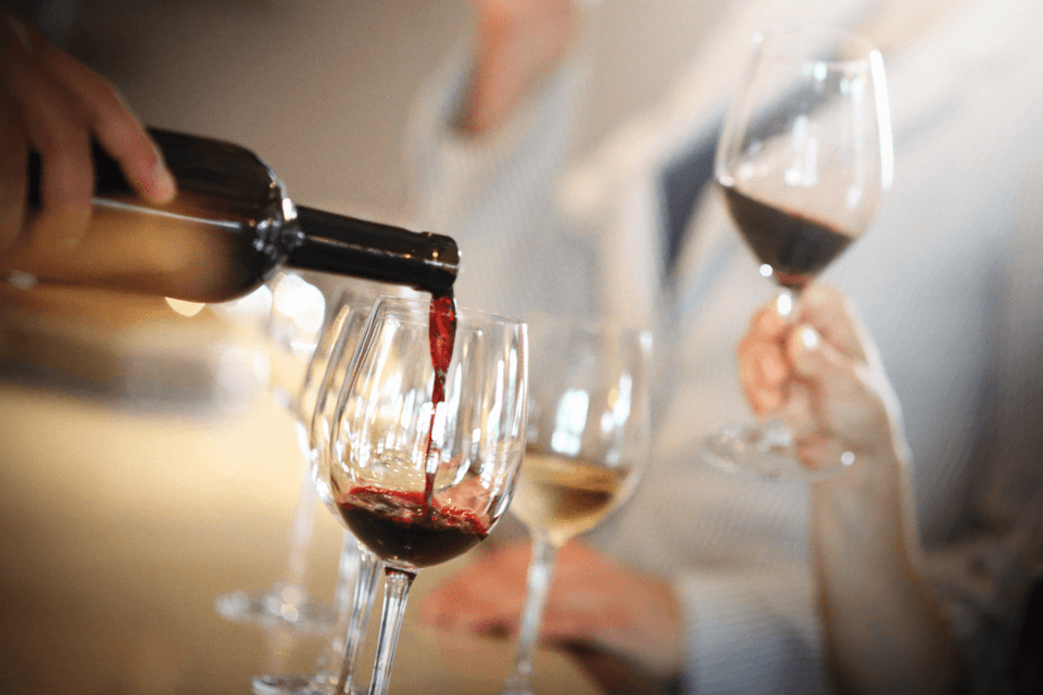 Primo piano di una mano che versa vino rosso da una bottiglia scura in un calice di vino trasparente durante una degustazione.