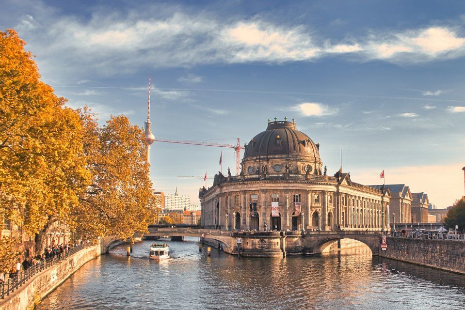 Un edificio a cupola sulla riva di un fiume, con alberi dalle foglie autunnali gialle e un'alta torre televisiva sullo sfondo.