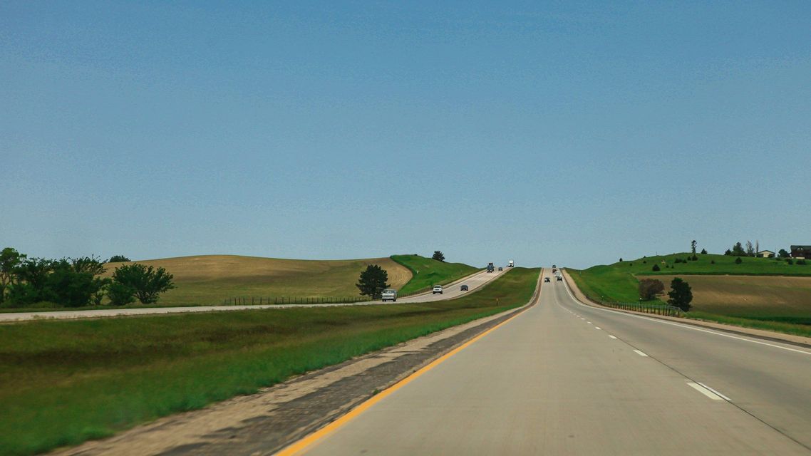 Un'autostrada attraversa un paesaggio di dolci colline verdi e marroni sotto un vasto cielo azzurro e limpido.