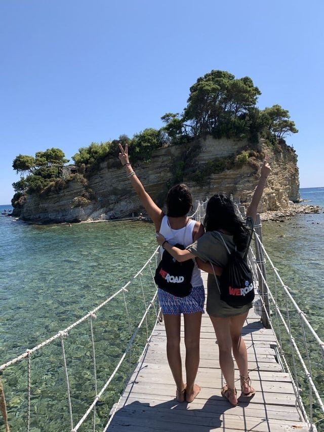 Zwei Frauen einer WeRoad-Gruppenreise, von hinten gesehen, stehen auf einer Seilbrücke über klarem Wasser und machen Friedenszeichen zu einer kleinen Insel.