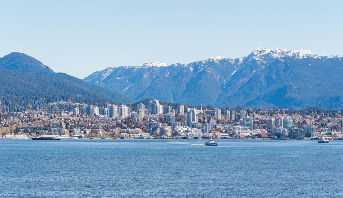 Uno skyline cittadino su uno specchio d'acqua, con grandi montagne innevate che si ergono sullo sfondo sotto un cielo sereno.