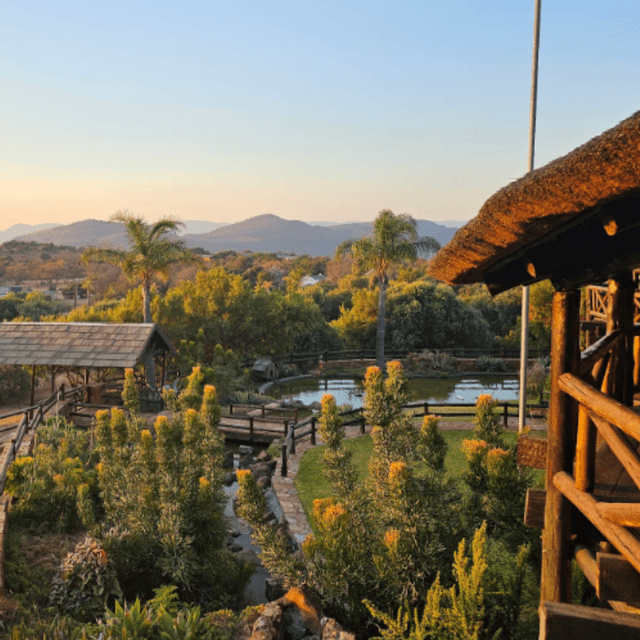 Un jardin luxuriant avec un étang et des sentiers en bois, vus depuis un pavillon au toit de chaume, avec des montagnes en arrière-plan au coucher du soleil.