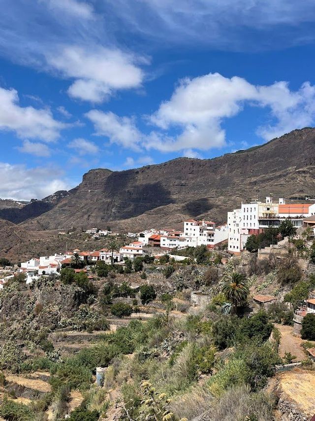 Un villaggio di edifici bianchi con tetti di tegole rosse, adagiato su un verde fianco di montagna terrazzato, sotto un cielo azzurro e nuvoloso.