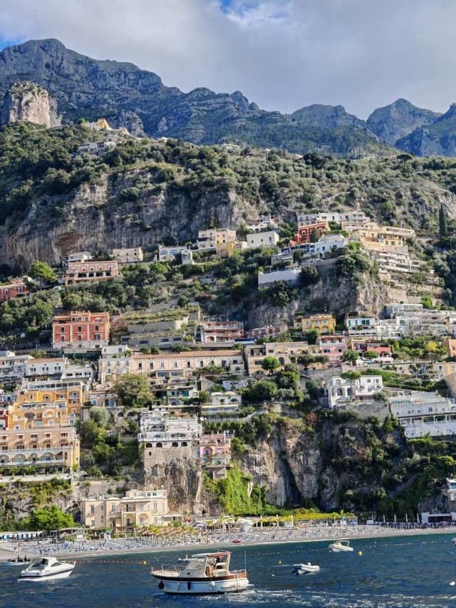 Una cittadina con edifici colorati è incastonata in una ripida scogliera verde, affacciata su una spiaggia con barche sull'acqua.