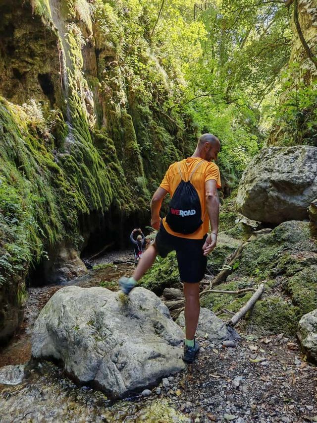Ein Mann mit einem WeRoad-Rucksack wandert durch eine enge, moosbewachsene Schlucht und steigt über einen großen Felsen in einem Bach.