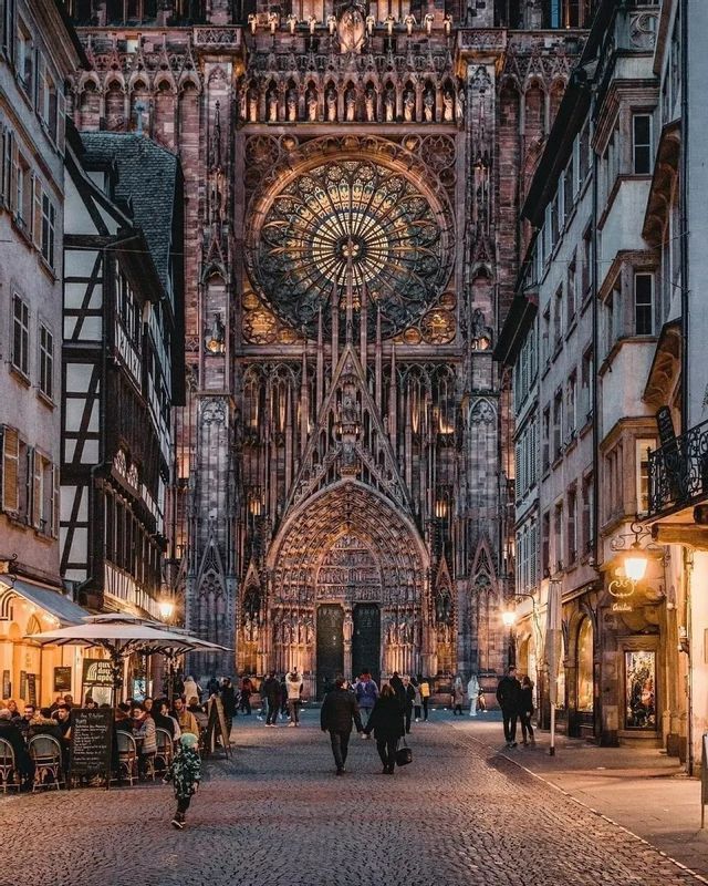 The illuminated facade of a large Gothic cathedral with a detailed rose window, seen from a narrow cobblestone street at dusk.