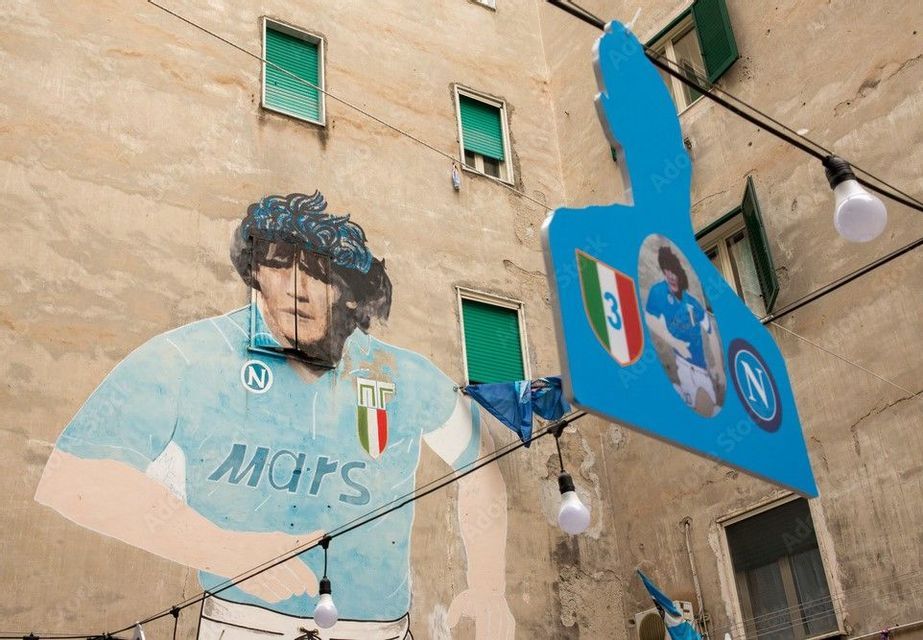 Un murale del calciatore Diego Maradona con una maglia azzurra su una facciata di un edificio, con luci pendenti e insegne davanti.