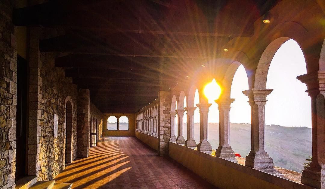 Il sole al tramonto splende attraverso gli archi di un colonnato in pietra, proiettando lunghe ombre dai pilastri su un pavimento piastrellato.