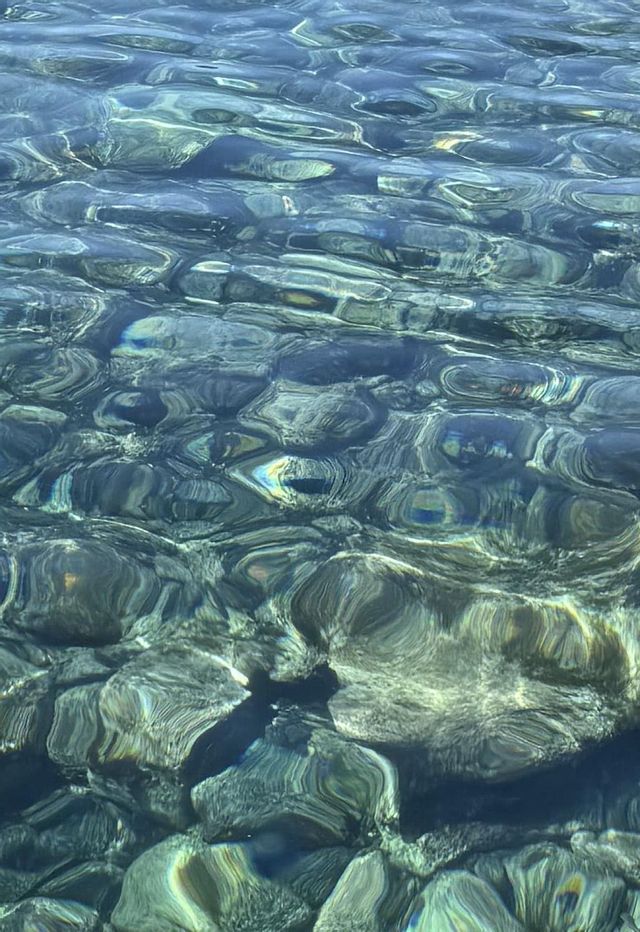 La luce del sole si riflette sulla superficie dell'acqua limpida e increspata, rivelando sul fondo pietre lisce e scure.