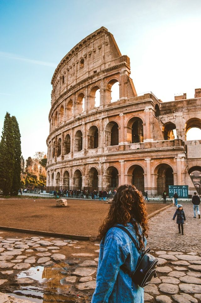 A woman with curly hair and a denim jacket stands on a cobblestone street, looking up at a large, ancient stone amphitheater under a clear sky.