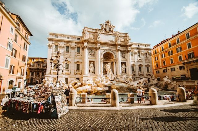 Una gran fuente ornamentada en una plaza empedrada, con turistas reunidos alrededor y un puesto de souvenirs en primer plano.
