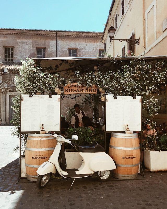 Un scooter blanc est garé dans une rue pavée, devant un stand de restaurant extérieur avec deux grands tonneaux en bois.