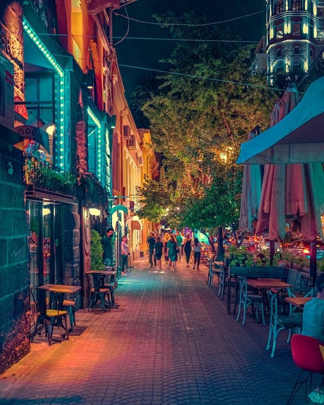 A narrow city street at night with people walking past outdoor cafes illuminated by colorful neon lights.