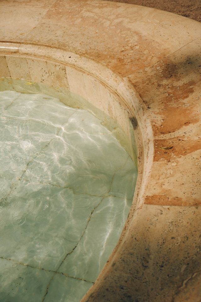 A close-up of a curved stone fountain edge with clear water inside, where sunlight creates reflections on the tiled bottom.