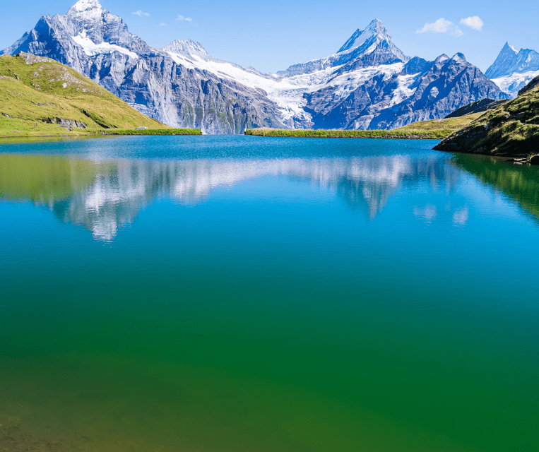 Snow-capped mountains reflecting in the still, blue-green water of an alpine lake under a clear sky.