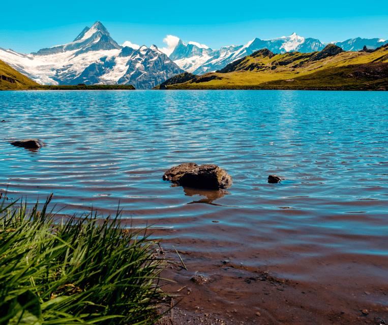 A low-angle view from a grassy shore of a clear alpine lake, with jagged, snow-capped mountains rising in the distance under a bright blue sky.
