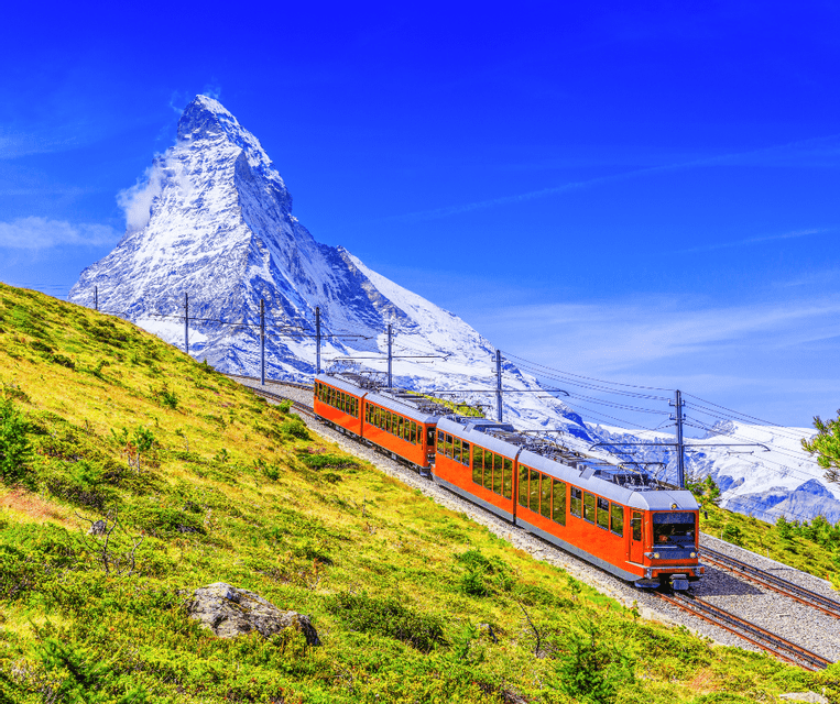 An orange train travels along a mountainside track with a snow-covered peak in the background under a clear blue sky.